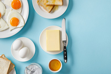 top view of butter, toasts, fresh and fried eggs on white plates, yogurt with chia seeds and bowl with honey on blue background