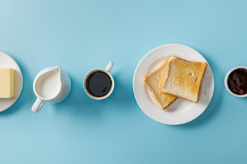 Top view of cup of coffee, milk, butter, jam and two toasts on white plate on blue background