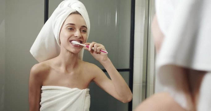 Pretty smiling woman with towel on head cleaning teeth with brush and having fun in the bethroom