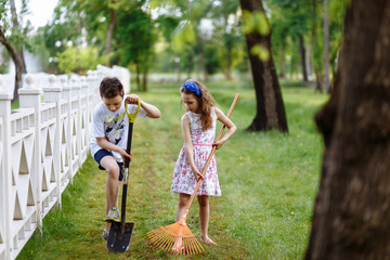 Fototapeta premium Cheerful little girl with rake and boy digging with shovel near the wooden fence in the park. Brother and sister works in garden. Childhood concept.