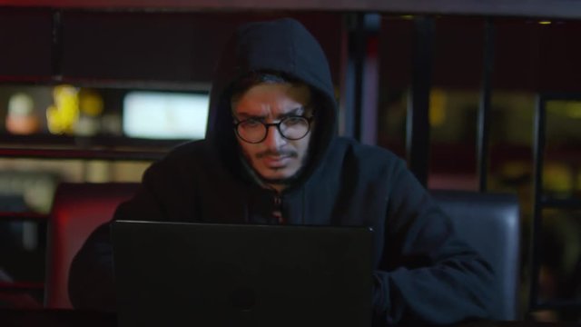 Zoom in of young Arab man in hoodie sitting at laptop and typing code on keyboard