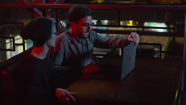 Medium shot of two hackers sitting down at caf&eacute; table, putting laptop on it and starting coding