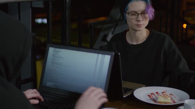 Tilt up of two hackers sitting at caf&eacute; table in front of each other, typing program code, eating dessert and drinking tea