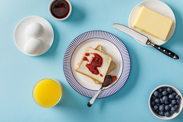 top view of toasts, bowl with jam, spoon and knife on plates, boiled eggs, butter, blueberries and orange juice on blue background © LIGHTFIELD STUDIOS
