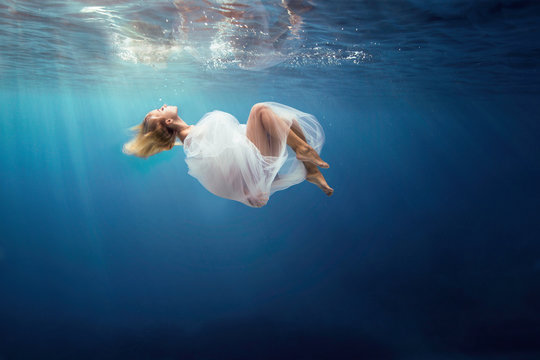 Blonde girl wrapped in fine white cloth, sank in blue deep water of ocean, against dark sea background.