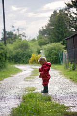 Sweet little toddler boy, blond child in red raincoat and blue boots, playing in the rain in muddy puddles, jumping and laughing