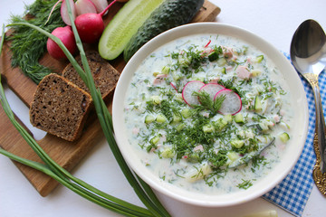 Okroshka is a traditional Russian summer cold soup in a ceramic bowl. Ingredients potatoes, radishes, cucumbers, dill, eggs, kefir yogurt, sausage. Summer yogurt cold soup on a wooden table. Selective