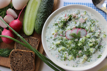 Okroshka is a traditional Russian summer cold soup in a ceramic bowl. Ingredients potatoes, radishes, cucumbers, dill, eggs, kefir yogurt, sausage. Summer yogurt cold soup on a wooden table. Selective