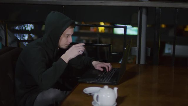 Tracking left of young hooded man sitting in caf&eacute; at laptop, typing on keyboard and drinking tea from tea cup