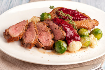 Roasted duck breast with vegetable garnish. The meat is sliced into portions and sprinkled with cranberry berry sauce. Large portion on a plate. Close up and horizontal view.