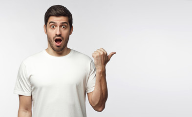 Young surprised man in white t-shirt looking at camera with open mouth, pointing right, copy space...