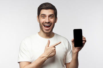 Excited screaming WOW young man in white t-shirt presenting smartphone and pointing with finger at...