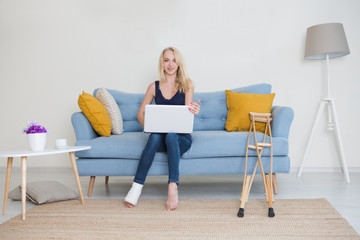 Young woman with  broken leg in cast sitting on sofa at home working on the laptop