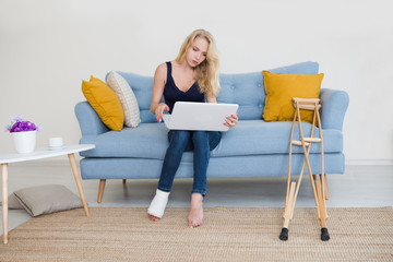 Young woman with  broken leg in cast sitting on sofa at home working on the laptop