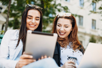 Portrait of two beautiful young girlfriends sitting on a bench looking on a tablet smiling against a building.