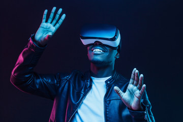 Young african man wearing virtual reality goggles with hands up, isolated on black background