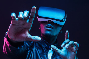 Young african man wearing virtual reality goggles with hands up, isolated on black background