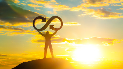 A silhouette of a man holds an infinity symbol on top of a mountain with a sunset background....
