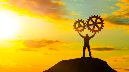 A man at the top of a mountain collects a puzzle from gears, against a dramatic sky in the sunset. Business concept idea. Innovation, cooperation, partnership, teamwork