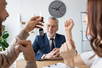 selective focus of happy car dealer looking at man holding car keys near cheerful woman