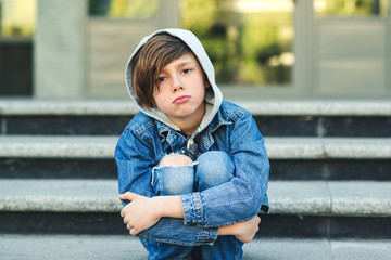 Sad boy is sitting on the stairs, before school. Alone unhappy child in city street. Bullying, depression, child protection or loneliness concept. Depressed teenage boy