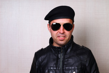 Cool rocker in black leather jacket and sunglasses on blurred wall background in studio. Emotion of...