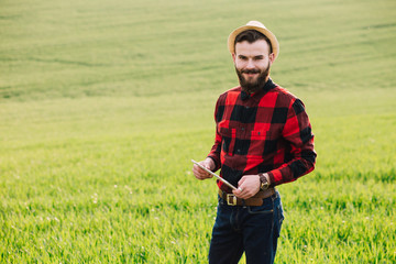 Naklejka na ściany i meble Young handsome bearded farmer with tablet standing in wheat field in early summer