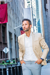 Young handsome European Man with beard traveling in New York City. Young guy wearing beige blaze, white shirt, gray pant, walking on narrow vintage street with banners, looking around, thinking.