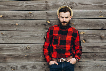 Obraz premium Portrait of a young stylish bearded man dressed up in jeans and a true worker black by red shirt on a wooden background.
