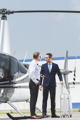 Pilot in formal wear and businessman with suitcase near helicopter