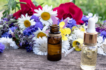 Essential oil in a glass bottle near wildflowers on wooden background.