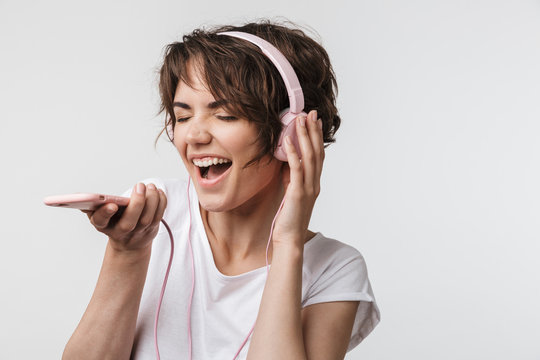 Image of joyous woman in basic t-shirt singing while listening to music with headphones