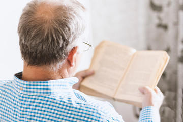 Middle-aged gray man with glasses in blue shirt reading book in room, soft focus