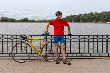 Athlete stands next to his road bike on waterfront of lake. Natural background.