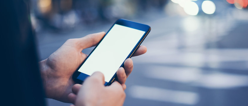 Closeup image of male hand holding smartphone with blank screen. Mockup ready for text message or content. Man's hands with cellphone. Empty display. Night street, bokeh light