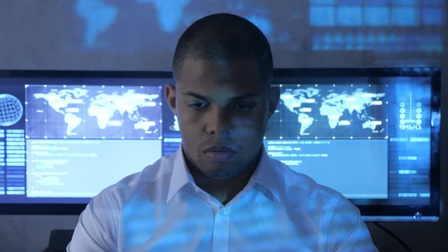 Portrait of African American man programmer in white shirt working on computer in security data center. Holographic code on his face