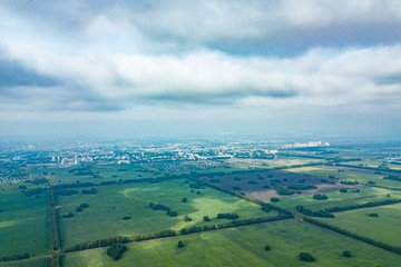 Aerial view of a green agricultural field and meadows with grass and plants separated by a country road in the middle and a strip of windproof trees on a sunny summer day with grayand white clouds