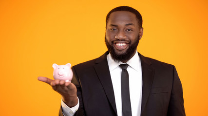 Joyful african-american man in suit putting coin in piggybank retirement savings