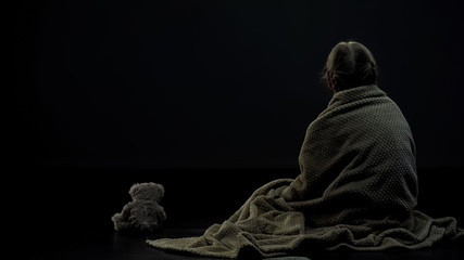 Little female kid in plaid sitting near teddy bear in dark room, loneliness