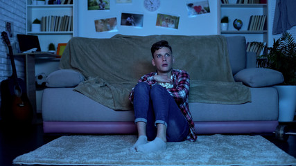 Obraz premium Shocked teenage boy watching horror movies on tv late at night, emotions