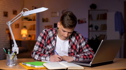 Teenager doing his homework, writing task into copybook, laptop on table