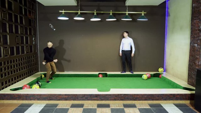 Two men playing billiards, hitting balls with legs, kicking it into holes on a floor billiard table. 4K