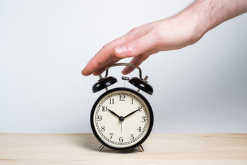 A man puts his hand on a retro black alarm clock on a wooden table on a white background. Good morning. Deadline. Not enough sleep