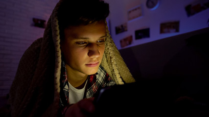 Tired teenager surfing internet on tab instead of sleeping, lying under blanket
