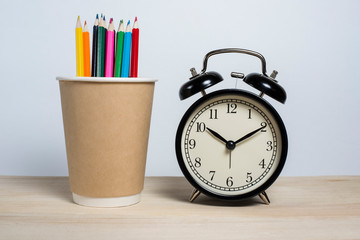 Black retro alarm clock next to colored pencils on a wooden table on a white background. Deadline. Good morning. Training at school and university.