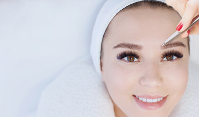 Beautiful young girl with long eyelashes tweezing her eyebrows in a beauty salon. Woman doing eyebrow permanent makeup correction . Microblading brow.