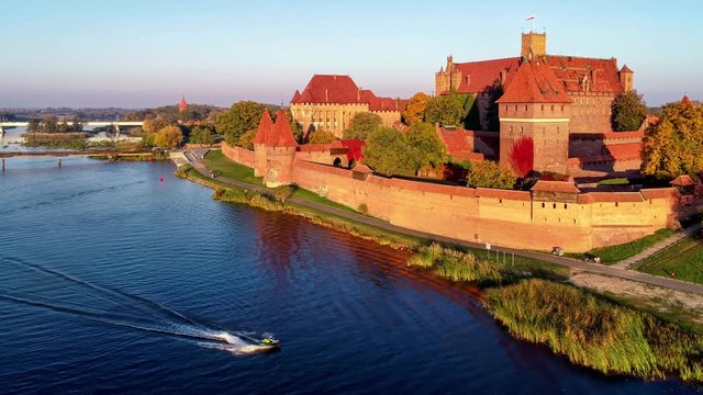Medieval Malbork (Marienburg) Castle in Poland, main fortress of the Teutonic Knights at the Nogat river and a water scooter. Aerial flyby 4K video in sunset light