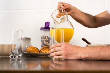 cropped view of man pouring orange juice in glass during breakfast in kitchen