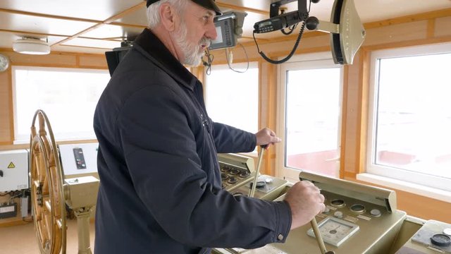 Bearded gray haired captain of ship in peaked cap and uniform at work in ship cabin operates the ship with two levers. Shipping concept.