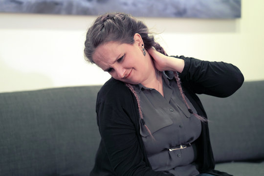 Woman suffering from neck pain at home on couch. A woman's sense of fatigue, exhausted, stressed. Woman massages her painful neck with her hands. The concept of body and health.